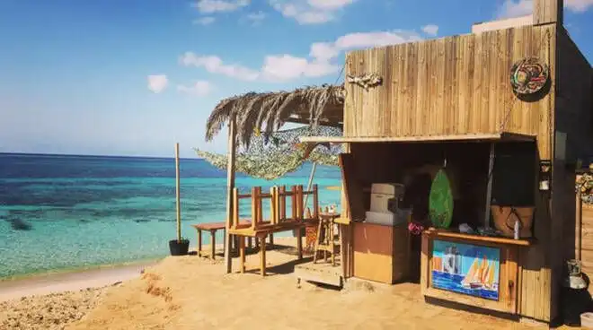 Un bar rustico di legno con tetto in paglia si trova su una spiaggia sabbiosa, con vista su un mare calmo e turchese sotto un cielo azzurro.