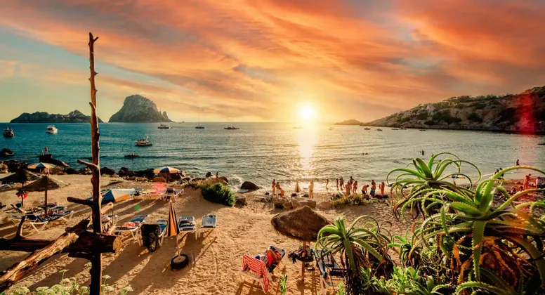 Un tramonto arancione vivido su una cala balneare piena di gente, con barche sull'acqua e una grande isola rocciosa in lontananza.