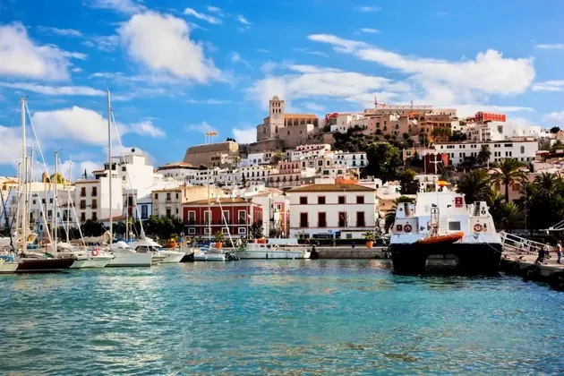 Un porto con barche a vela e yacht ormeggiati, davanti a una cittadina collinare con edifici bianchi sotto un cielo azzurro.