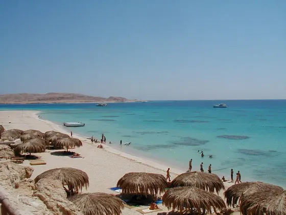 Blick von oben auf einen tropischen Strand mit Strohschirmen, Badenden im türkisfarbenen Wasser und Booten in der Ferne.