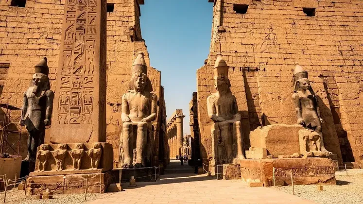 Große Steinstatuen von Pharaonen flankieren einen sonnendurchfluteten Korridor, der zu einem altägyptischen Tempel führt, dessen hohe Mauern mit Hieroglyphen bedeckt sind.