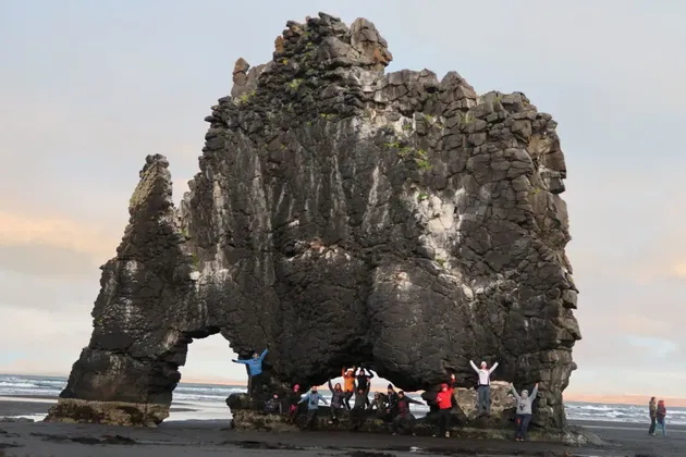 Un gruppo WeRoad in viaggio posa per una foto sotto una grande formazione rocciosa ad arco su una spiaggia di sabbia scura.