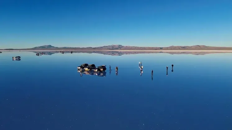 Un viaggio di gruppo WeRoad su una sconfinata distesa salina riflettente con veicoli 4x4, creando uno specchio perfetto del cielo azzurro e delle montagne.