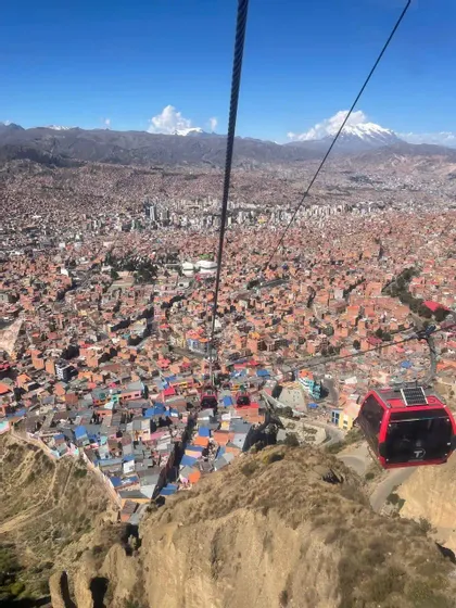 Una funivia rossa sorvola una vasta città in una valle, con montagne innevate sotto un cielo azzurro e limpido.