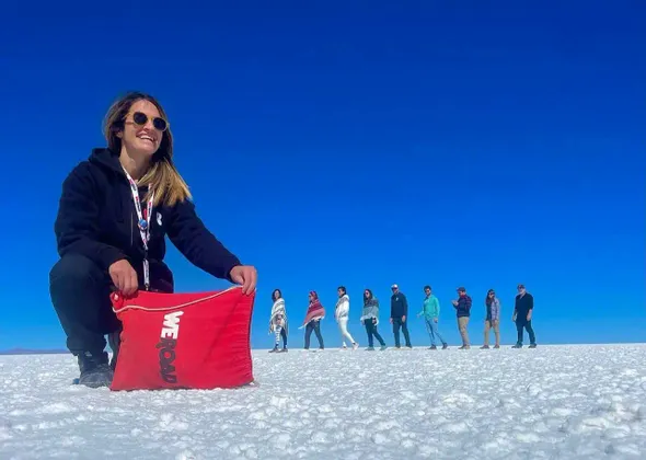 Una donna si accovaccia tenendo una borsa WeRoad rossa su una vasta distesa di sale bianca, con il gruppo di viaggio WeRoad allineato sullo sfondo sotto un cielo azzurro e limpido.