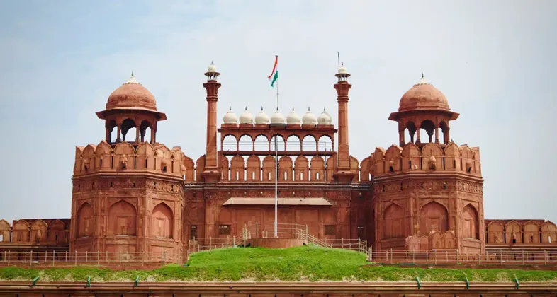 La bandiera dell'India sventola sopra i bastioni e le cupole di arenaria rossa di una grande fortezza simmetrica sotto un cielo coperto.