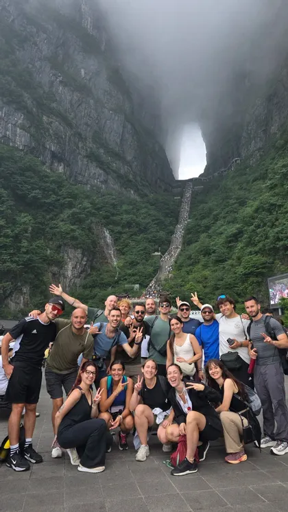 Un gruppo WeRoad in viaggio si fa fotografare ai piedi di una montagna ripida e verde, con un grande arco naturale e una lunga scalinata.