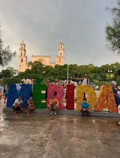 Un groupe WeRoad en voyage pose pour une photo devant de grandes lettres colorées formant 'MERIDA' sur une place avec une cathédrale en arrière-plan.