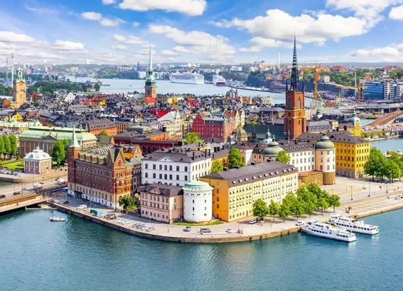 Vista aérea de una ciudad europea histórica con edificios coloridos y agujas a lo largo de un paseo marítimo bajo un cielo azul.