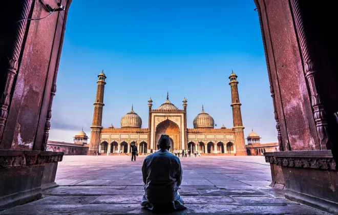 Ein von hinten betrachteter Mann kniet in einem steinernen Torbogen und blickt auf eine große Moschee mit Minaretten jenseits eines Hofes.