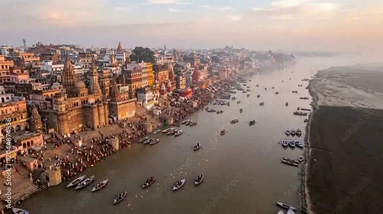 Eine Luftaufnahme einer geschäftigen Stadt an einem Fluss, mit zahlreichen Booten auf dem Wasser und Menschenmengen auf den steinernen Ghats bei Sonnenuntergang.