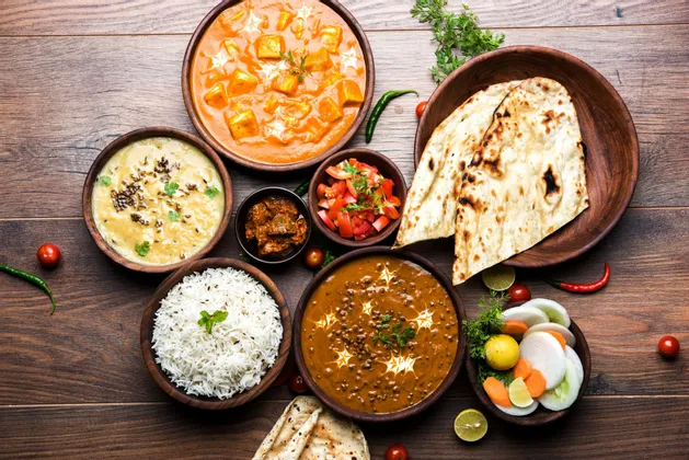 Eine Draufsicht auf ein traditionelles indisches Gericht mit verschiedenen Currys, Reis und Naan-Brot, serviert in Schüsseln auf einer Holzoberfläche.