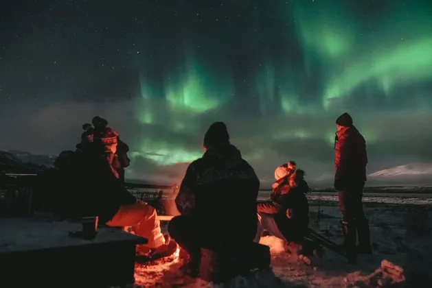 Eine WeRoad-Gruppe sitzt nachts in einer verschneiten Landschaft am Lagerfeuer und beobachtet die grünen Nordlichter am Sternenhimmel.