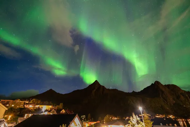 Die Nordlichter leuchten grün und lila an einem sternenklaren Nachthimmel über der dunklen Silhouette von Bergen und einem beleuchteten Dorf.