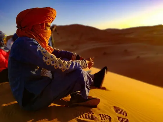 Una persona en un viaje grupal de WeRoad, con un turbante naranja, se sienta en una duna de arena observando el atardecer en el desierto.
