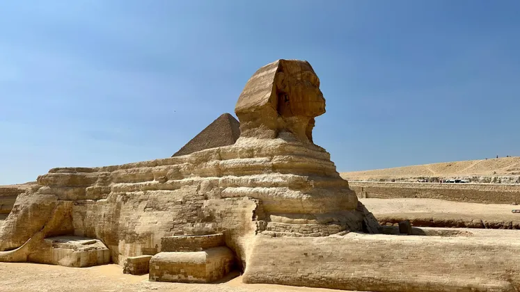 Seitenprofil eines massiven Steinsphingenmonuments in einer Wüstenlandschaft mit einer Pyramide im Hintergrund