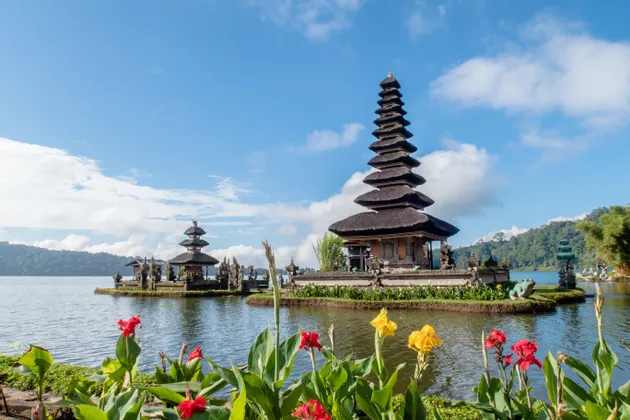 Zwei traditionelle Tempel mit gestuften Dächern liegen an einem See, mit roten und gelben Blumen im Vordergrund unter einem teilweise bewölkten Himmel.