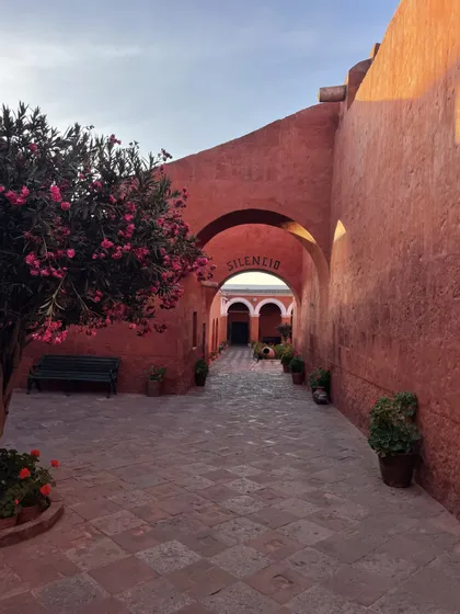 Cortile rosso terracotta con pavimentazione in pietra, albero in fiore rosa e un arco contrassegnato dalla parola SILENCIO.