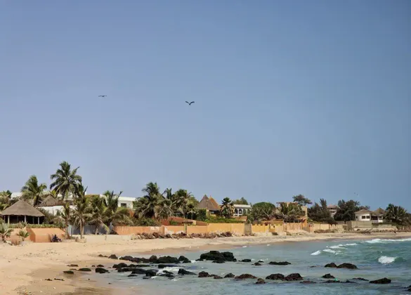 Ein Küstendorf mit Palmen und traditionellen Gebäuden liegt an einem Sandstrand, neben einer felsigen Küste, unter klarem blauem Himmel.