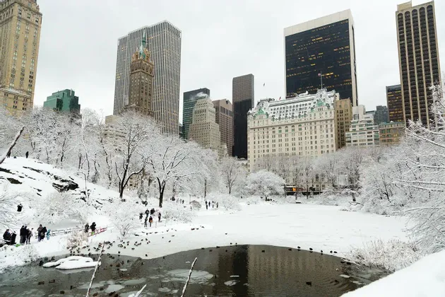 Ein schneebedeckter Park mit einem teilweise zugefrorenen Teich und spazierenden Menschen, vor der Kulisse von Stadtwolkenkratzern.