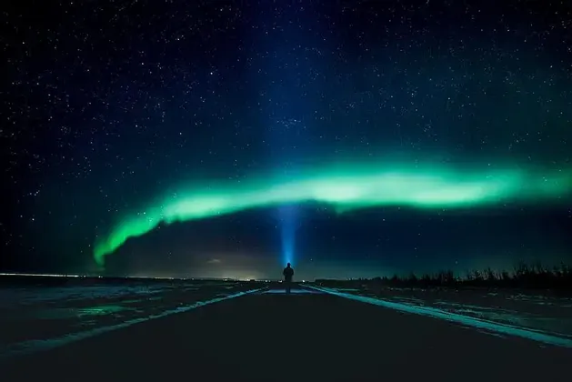 Una persona si trova su una strada buia di notte, puntando una luce verso l'aurora boreale verde nel cielo stellato.