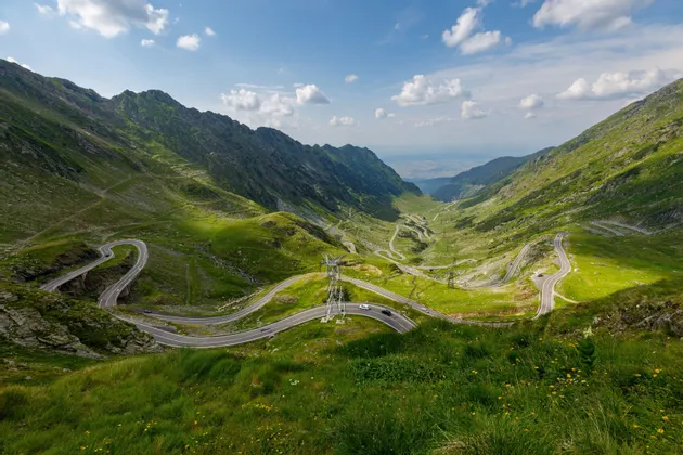 Una strada tortuosa con auto attraversa una rigogliosa valle di montagna verde sotto un cielo azzurro con alcune nuvole.