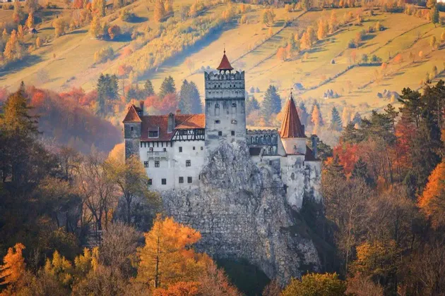 Un castello in stile medievale con tetti in tegole rosse, arroccato su una scogliera rocciosa, circondato da una fitta foresta con colori autunnali.