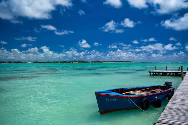 Una barca blu è ormeggiata a un molo di legno in acqua calma e turchese sotto un cielo blu con nuvole bianche.