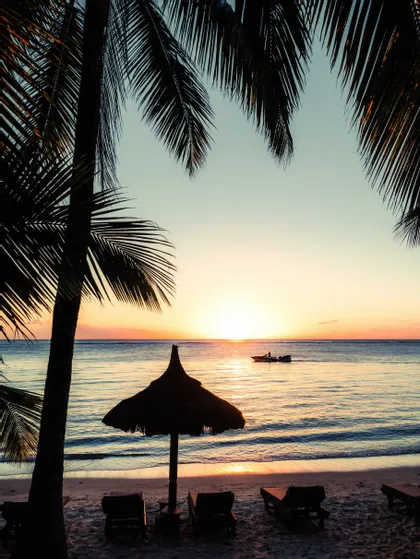 Silhouette di palme e di un ombrellone di paglia su una spiaggia sabbiosa durante un tramonto vivace sull'oceano, con una piccola barca in lontananza.