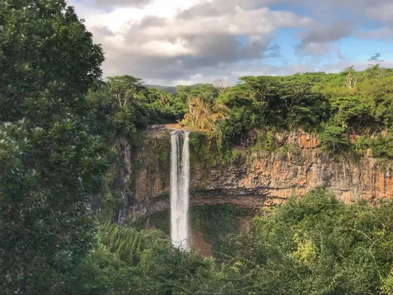 Una cascata imponente scende da una scogliera rocciosa, circondata da una giungla verde lussureggiante, sotto un cielo parzialmente nuvoloso.