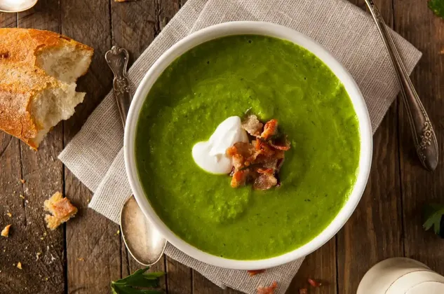 Eine Schale grüner Suppe, garniert mit Sahne und Speckwürfeln, steht auf einem Holztisch neben einem Stück knusprigem Brot und einem Löffel.
