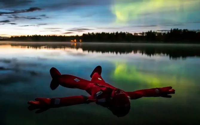 Eine Person in einem roten Überlebensanzug treibt in einem ruhigen See auf dem Rücken, während sich die grünen Nordlichter in der Dämmerung auf dem Wasser spiegeln.