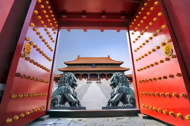 Große, offene rote Tore mit goldenen Knäufen umrahmen einen traditionellen chinesischen Palast, der von zwei bronzenen Löwenstatuen bewacht wird.