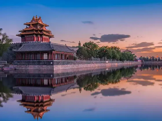Una torre de esquina tradicional china sobre un muro de piedra se refleja en las tranquilas aguas de un foso al atardecer.