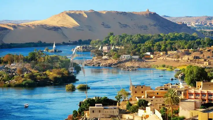 Un amplio río azul con veleros, bordeado por un pueblo con palmeras y una gran duna de arena dorada.