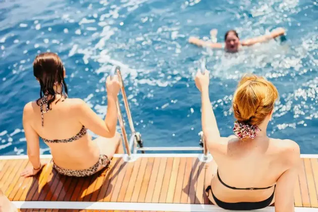 Zwei Frauen einer WeRoad-Gruppenreise sitzen auf dem Holzdeck eines Bootes und beobachten eine andere Frau, die im blauen Meer schwimmt.