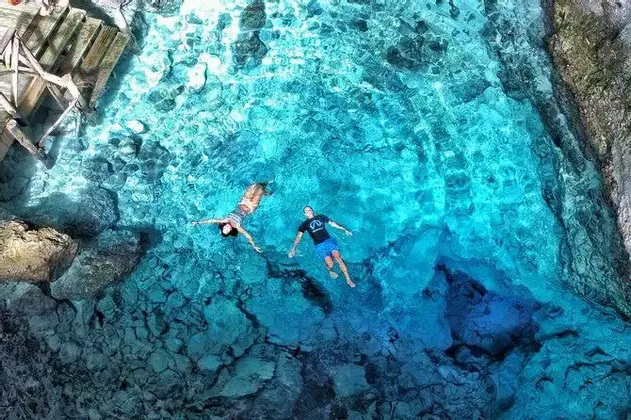 Vista aerea di un uomo e una donna che galleggiano sulla schiena in acqua limpida e turchese sopra un fondale roccioso.