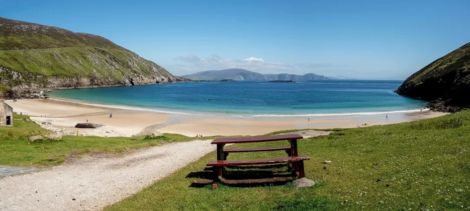 Una panchina di legno su una collina erbosa si affaccia su una spiaggia sabbiosa appartata e una baia turchese tra due grandi scogliere.