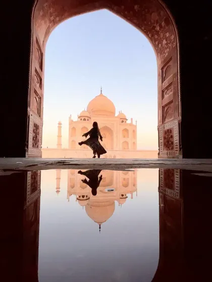 Una persona in silhouette posa davanti al Taj Mahal, ripresa attraverso un arco e con il suo riflesso in una pozzanghera in primo piano.