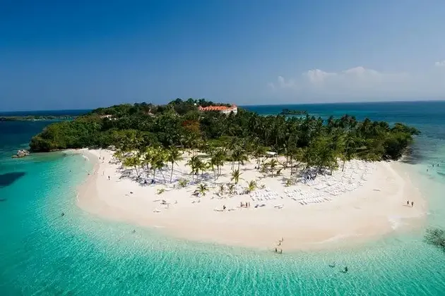 Una vista aerea di un'isola tropicale con una spiaggia di sabbia bianca, palme e acque cristalline color turchese dove le persone nuotano e si rilassano.