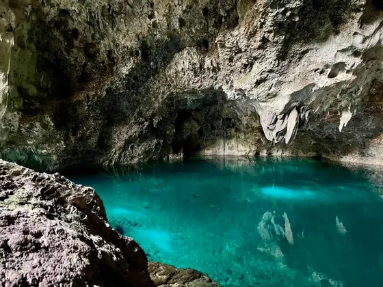 Una piscina naturale sotterranea di acqua limpida e turchese che brilla dall'interno di una grotta buia e rocciosa con stalattiti sul soffitto.