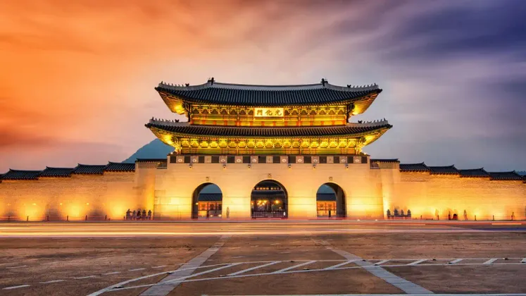 Ein traditionelles asiatisches Torhaus und seine Steinmauer sind in der Dämmerung hell erleuchtet, mit langen Lichtspuren von Fahrzeugen im Vordergrund.