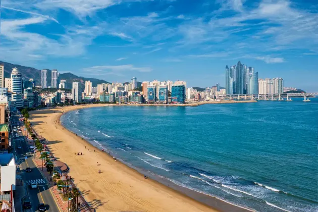 Vista aérea de una amplia playa de arena que serpentea junto a una ciudad moderna con rascacielos, con las aguas azules del océano bañando la orilla bajo un cielo soleado.