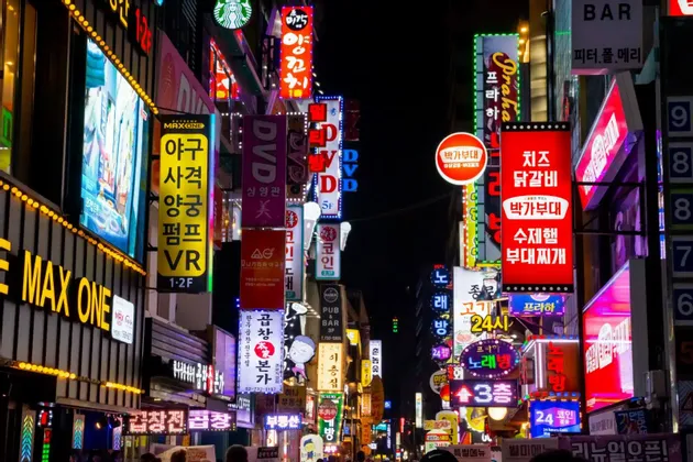 Eine enge Gasse ist nachts hell erleuchtet von einer dichten Wand farbenfroher Neonschilder, meist in koreanischer Schrift.