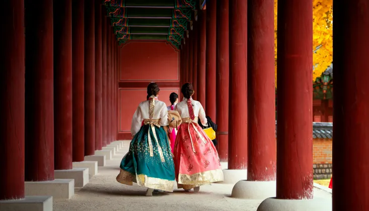 Drei Frauen, von hinten gesehen, tragen farbenfrohe traditionelle Hanbok-Kleider und gehen durch einen Korridor mit großen roten Säulen.