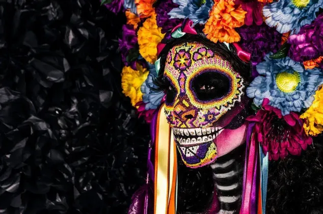 Una persona di profilo con trucco colorato da teschio di zucchero Calavera e un elaborato copricapo di fiori multicolori su sfondo nero.