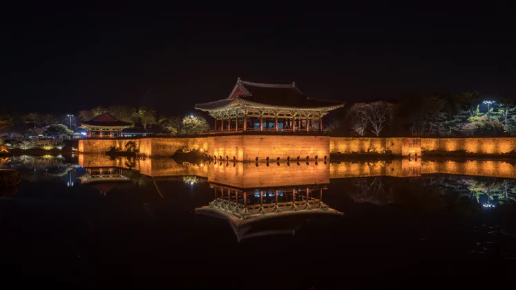 Ein traditioneller Pavillon und eine Steinmauer, nachts beleuchtet, werfen eine perfekte Spiegelung auf das ruhige Wasser darunter.