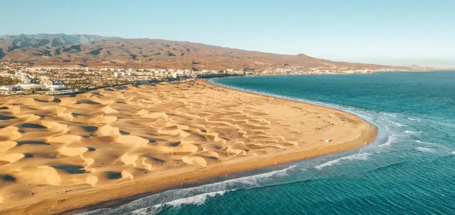 Une vue aérienne de vastes dunes de sable rencontrant l'océan bleu, avec une ville côtière et des montagnes en arrière-plan sous un ciel clair.
