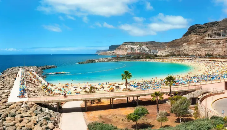 Vue aérienne d'une plage bondée dans une crique à l'eau turquoise, entourée de falaises sous un ciel bleu éclatant.