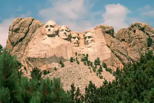 Il monumento del Monte Rushmore scolpito in una montagna di granito, con pini verdi in primo piano e un cielo azzurro sopra.
